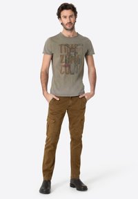 Olive green graphic t-shirt with text, paired with brown cargo pants featuring multiple pockets and a slim fit. Black shoes complete the outfit.