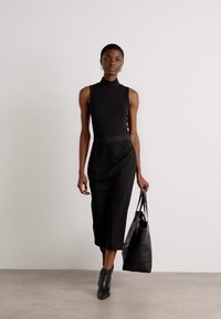 Top noir sans manches avec un col montant, associé à une jupe midi noire. Le modèle porte un sac fourre-tout noir texturé et des bottines noires.