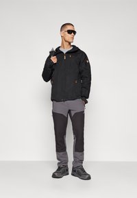 Zwarte waterdichte jas met capuchon en oranje accenten, gecombineerd met grijs en zwarte meerpaneelbroeken en stevige zwarte wandelschoenen.