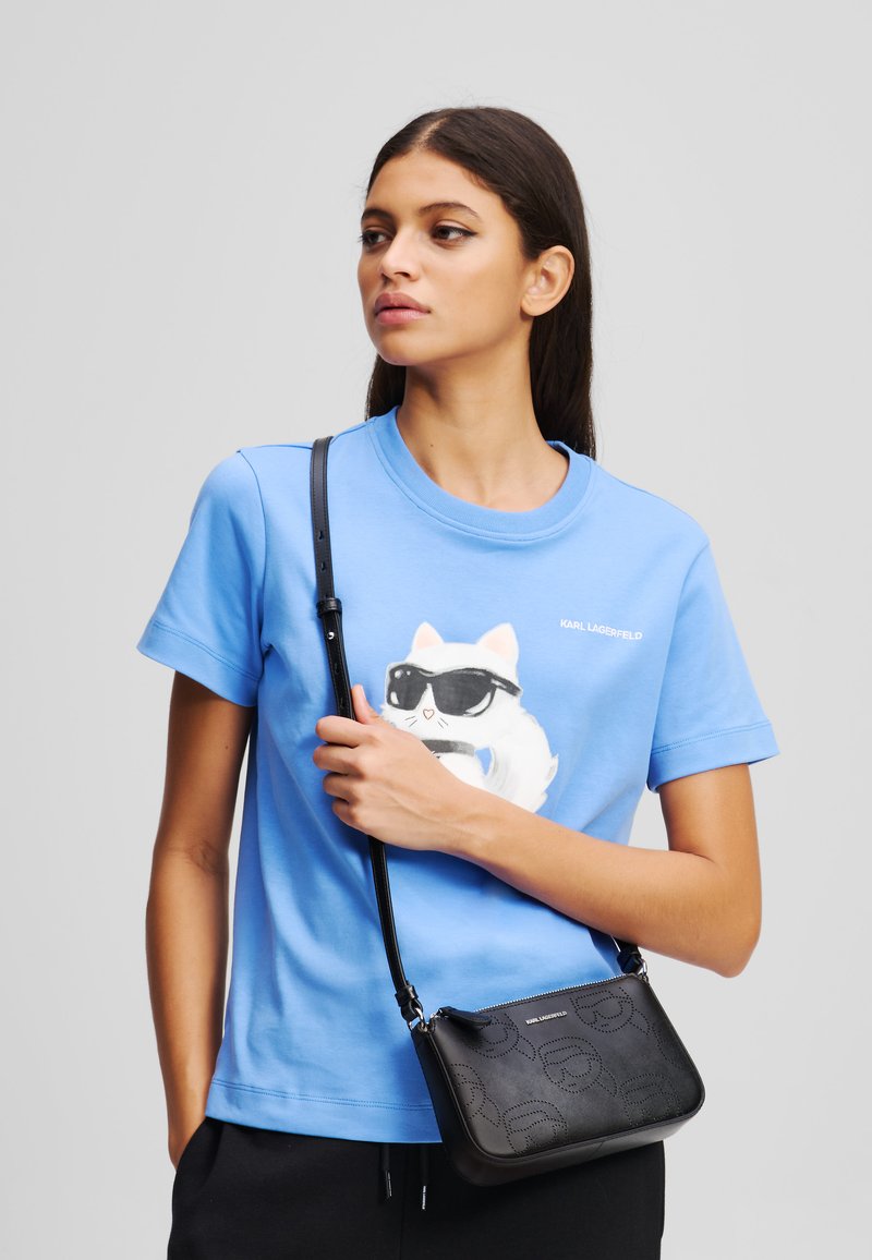 T-shirt di cotone azzurro chiaro con una grafica di un gatto che indossa occhiali da sole; abbinata a una piccola borsa a tracolla nera con design del logo in rilievo.