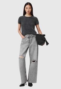 Grijze trui met korte mouwen en een zachte textuur, gecombineerd met oversized lichtgrijze gescheurde jeans. Het model houdt een zwarte tas met strikken vast.