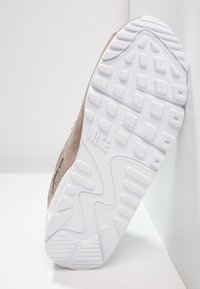 Teniska Nike s béžovým svrškem z broušené kůže, bílou gumovou podrážkou s texturovaným vzorem a značkovým označením, představující unikátní design podrážky.