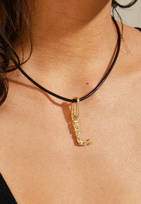 Gul halsbandshänge format som bokstaven "L" fäst vid ett svart läderhalsband. Närbild visar texturen och designens detaljer.