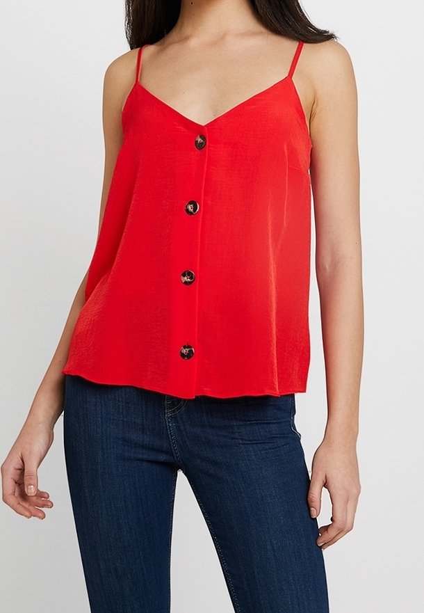 Femme portant un top rouge sans manches boutonné avec fines bretelles et un jean bleu foncé, debout devant un fond clair uni.