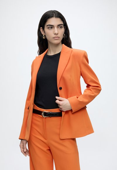 Femme aux longs cheveux foncés portant des boucles d'oreilles en or, un costume orange, une chemise noire et une ceinture noire avec une boucle dorée, posant sur un fond blanc.