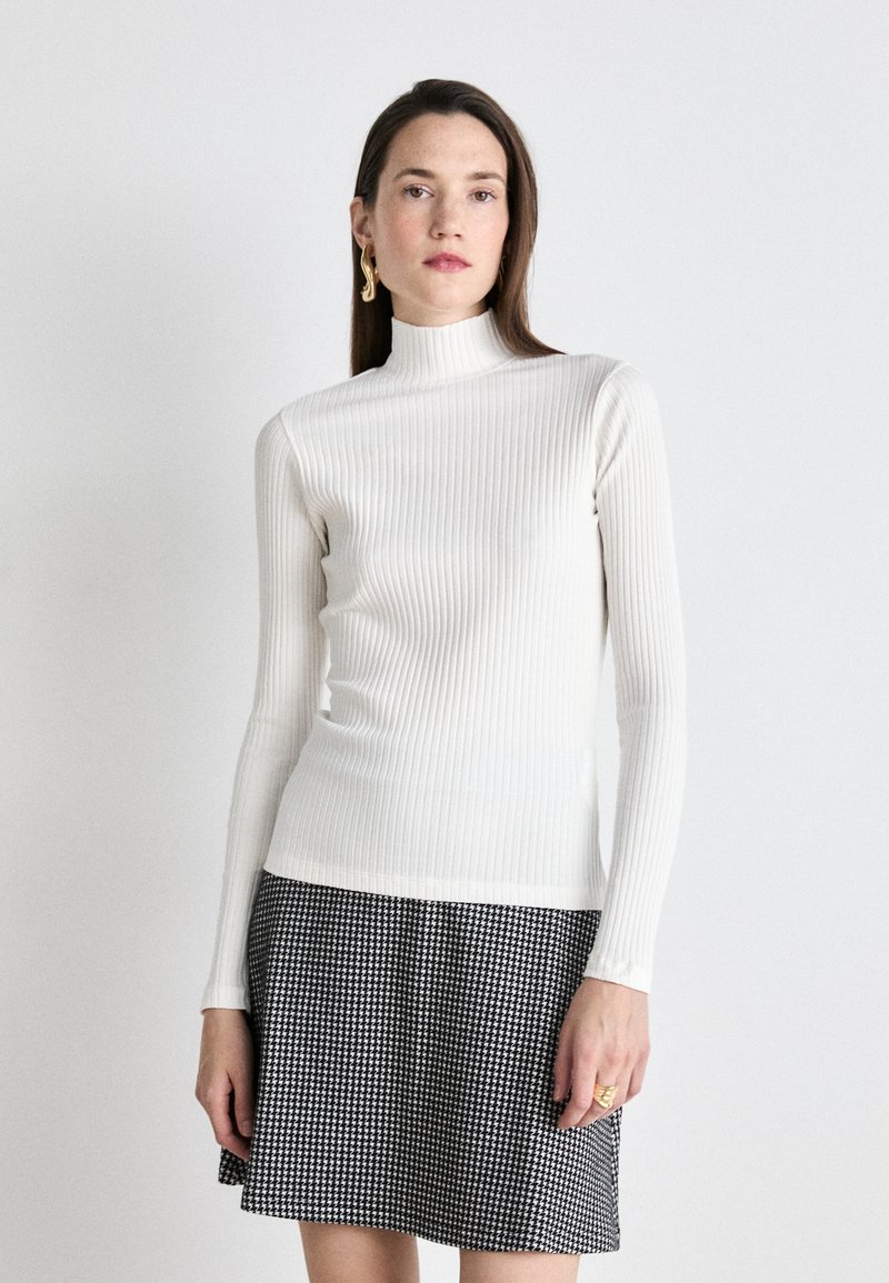 Maglione a collo alto bianco a coste con maniche lunghe abbinato a una gonna a quadretti bianca e nera, caratterizzata da una texture delicata e da un design aderente.