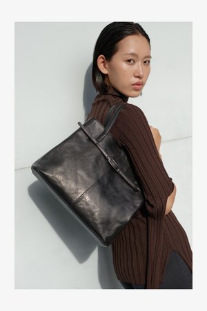 Ung kvinne iført en brun ribbestrikket genser bærer en stor svart lærveske over skulderen mot en ensfarget lys bakgrunn.