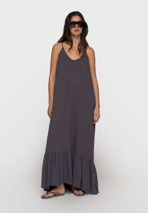 Donna che indossa un vestito lungo senza maniche grigio scuro con orlo arricciato, occhiali da sole neri oversize e infradito grigie, in piedi davanti a uno sfondo semplice.