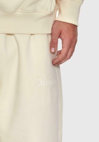 Hand, die auf schweißfarbenen Sweatpants mit dezentem "Dropöse"-Textstickerei ruht, gepaart mit dem Ärmel eines cremefarbenen Sweatshirts.