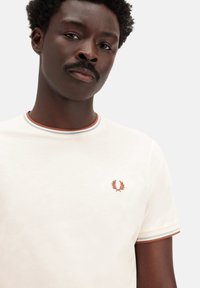 Camiseta de algodón blanca con cuello acanalado que presenta rayas azules y marrones. Pequeño logotipo bordado en el centro del pecho. Mangas cortas.