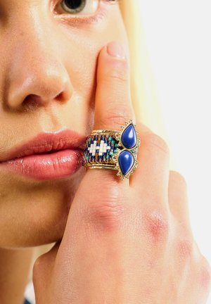 Mehrfarbiger Ring mit geflochtenem Band und Perlen sowie zwei blauen Tropfensteinen, gefasst in einem goldfarbenen Rahmen mit filigranen Details.