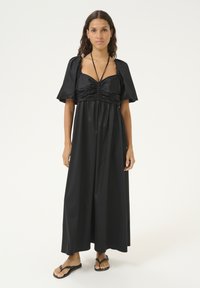 Sort satin maxikjole med korte puffærmer, rummelig bodice og slanke stropper. Modellen har simpelthen sorte flip-flops på.