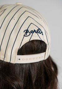 Hvid baseballkasket med marineblå striber, med "Berlin" broderet i kursiv og en justerbar plastrem bagpå.
