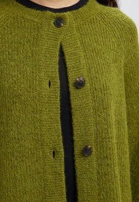 Cárdigan de punto verde con una textura acanalada, que presenta un escote redondo, tres botones oscuros y un pequeño detalle de agujero cerca del centro.