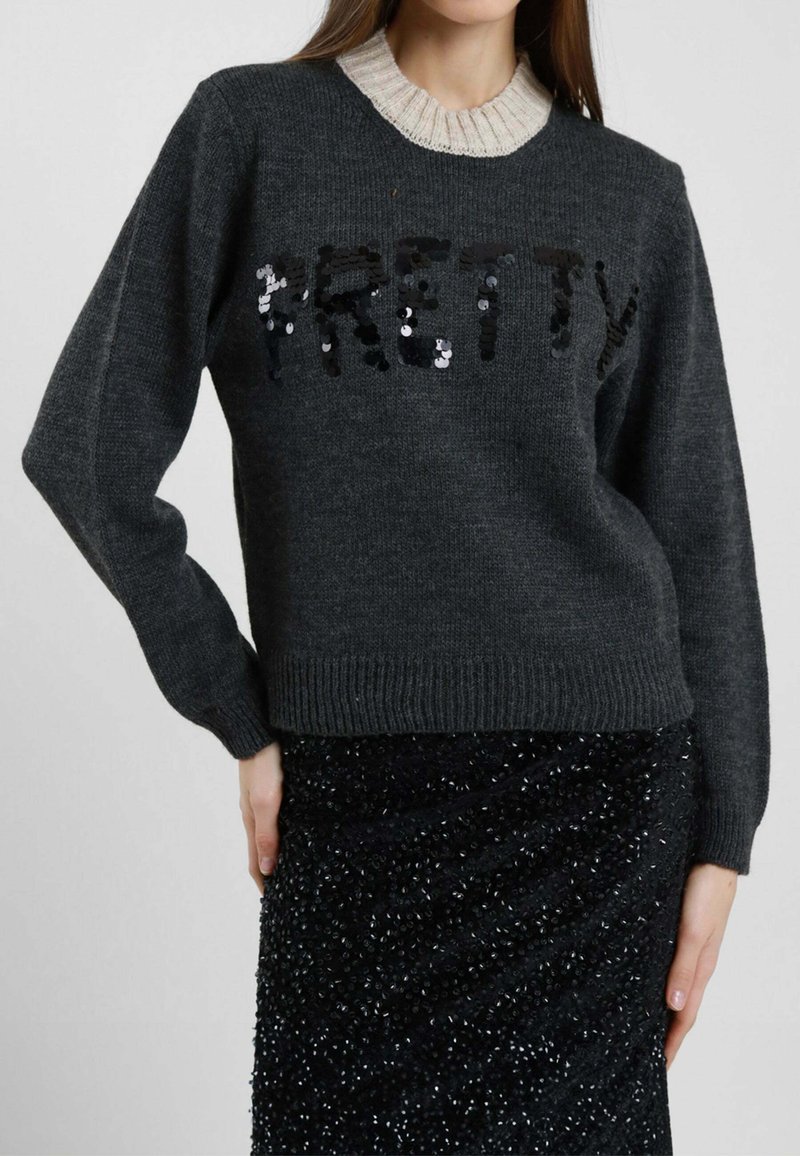 Donna che indossa un maglione grigio scuro con la scritta "PRETTY" in paillettes nere, sovrapposto a un colletto beige lavorato a maglia, abbinato a una gonna nera con paillettes.