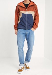 Windbreaker met kleurvlakken in brandoranje, beige en marineblauw, met ritssluiting, verstelbare capuchon en voorzakken; gecombineerd met lichtblauwe spijkerbroek en bruine sneakers.