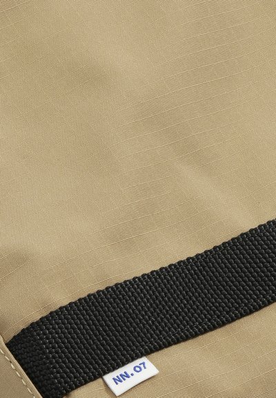 Tissu beige texturé avec une sangle noire tissée horizontale et une petite étiquette blanche portant l'inscription "NN.07" près de la sangle.
