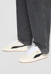 Sneakers Puma z zamszu w białym kolorze, z czarnym paskiem po boku i gumową podeszwą. Połączone z luźnymi szarymi spodniami, eksponującymi obuwie.