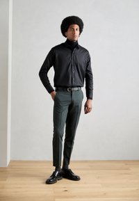 Camisa preta de botão, calças ajustadas verde-escuras e sapatos pretos de atar. Tecido suave, design elegante e um corte moderno.