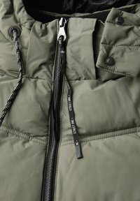 Khaki pufferjack met hoge kraag. Voorzien van een rits, drukknoopsluiting en een merknaam trekkoord. Gequilt textuur over het geheel.