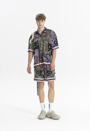 Joven de pie con camisa de manga corta con patrón de cachemira multicolor y pantalones cortos a juego, con calcetines blancos y zapatos beige texturizados sin cordones.