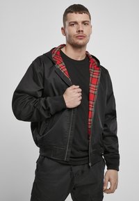 Zwarte jas gemaakt van soepel materiaal met een rode geruite binnenvoering. Beschikt over een capuchon, ritssluiting en zijzakken. Draagt over een zwart shirt.