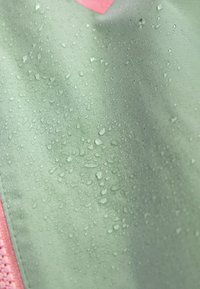 Des gouttes d'eau perlent sur un tissu vert clair avec un bord de fermeture éclair rose visible, indiquant un matériau résistant à l'eau.