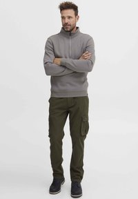 Felpa grigia con zip e alto collo, abbinata a pantaloni cargo verde oliva con tasche laterali e una vestibilità comoda. Gli stivali neri completano il look.