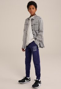 Siva denim jakna preko bele srajce, u kombinaciji s mornarskim trenirkama koje imaju logo. Crne atletske cipele s belim akcentima.