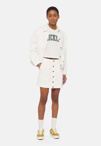 Weiße Denim-Jacke über einem weißen, kurz geschnittenen T-Shirt mit grünem Text, kombiniert mit einem weißen Rock mit Knopfleiste und gelben Wildlederschuhen.