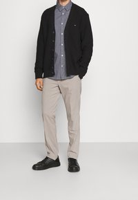 Cardigan nero a maglia con bottoni sopra una camicia blu a fantasia, abbinato a pantaloni beige e scarpe nere, il tutto su uno sfondo neutro.