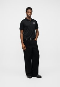 Νεαρός άνδρας στέκεται με το ένα χέρι στην τσέπη, φοράει μαύρο πόλο με μικρό λευκό λογότυπο, φαρδιά μαύρα παντελόνια, μαύρη ζώνη και μαύρα παπούτσια.