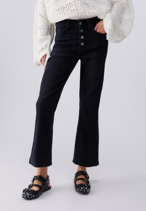 Personne portant un jean noir taille haute à boutons, un pull blanc texturé et des chaussures Mary Jane noires cloutées, debout devant un fond uni.