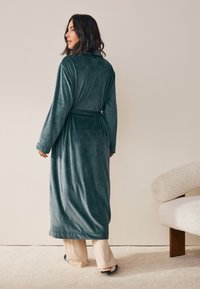 Dlhý tmavozelený župan z jemného zamatového materiálu s dlhými rukávmi a opaskom v páse. Lem siaha pod kolená.