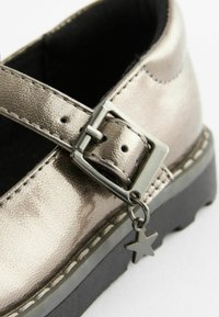 Metallisch silberner Mary Jane Schuh mit Schnalle und Sternenanhänger, mit strukturiertem Obermaterial, kontrastierender schwarzer Sohle und sichtbaren Nähdetails.