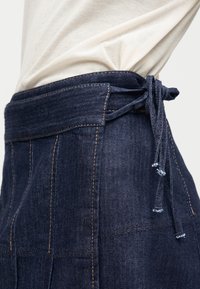 Κοντινή λήψη σκούρας μπλε τζιν φούστας με ορατές ραφές και δεμένο ζωνάρι, συνδυασμένη με ανοιχτό μπεζ πουκάμισο μέσα από τη φούστα.