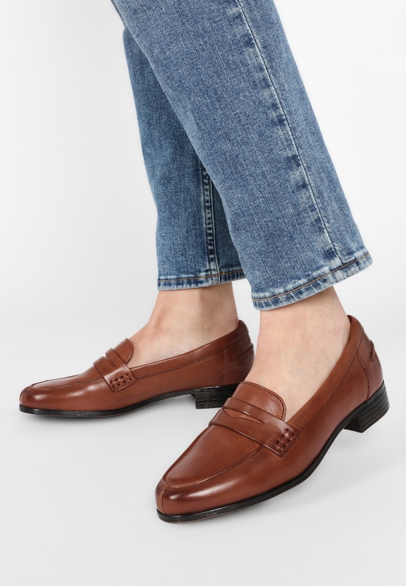 Mocassins en cuir marron avec un petit talon, présentant un détail cousu et une texture lisse. Portés avec un jean court bleu clair.