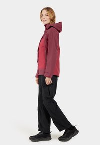 Veste à capuche bordeaux avec une poche zippée et des poignets élastiques, associée à un pantalon cargo noir et des chaussures de sport noires, se tenant sur un fond uni.