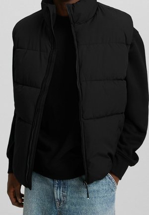 Hombre con chaleco acolchado negro sobre sudadera negra y vaqueros azul claro, de pie con una mano en el bolsillo contra un fondo liso.