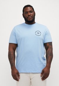 T-shirt di cotone blu chiaro con maniche corte, scollatura rotonda e un logo grafico circolare nero sulla parte sinistra del petto.