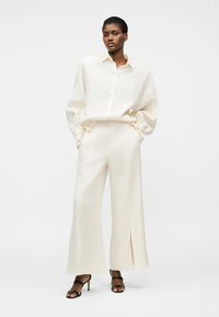 Nainen, joka pukeutunut löysään valkoiseen napitettuun paitaan ja leveälahkeisiin housuihin, joissa on etuslitsit, yhdistettynä mustiin korkeakorkoisiin sandaaleihin, seisoo valkoista taustaa vasten.