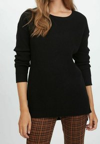 Schwarzer gerippter Strickpullover mit rundem Halsausschnitt und langen Ärmeln, der einen leicht verlängerten Saum hat. Kombiniert mit braunen Hahnentrittmuster-Hosen.