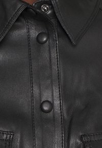 Camicia nera di pelle con pattina frontale e due bottoni a pressione, colletto appuntito e dettagli di cucitura visibili sulle tasche.
