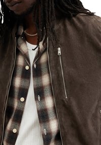 Brun ruskinds bomberjakke med en lynlås lomme, lagdelt over en ternet flannel skjorte med knapper og en hvid ribbestrikket sweater.