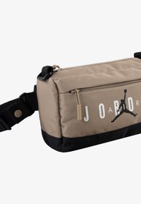Sac de bandoulière en tissu beige avec des accents noirs, de forme rectangulaire, compartiments zippés et logo Jordan imprimé en blanc.