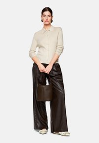 Beige gestrickte Strickjacke mit Kragen und Knöpfen auf der Vorderseite. Kombiniert mit hoch taillierten, braunen, aus Kunstleder gefertigten weit geschnittenen Hosen und einer dunkelbraunen Handtasche.