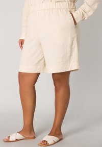 Beige linnen shorts met een elastische tailleband en zijzakken, met een ontspannen pasvorm en een licht gestructureerde stof. Gepaard met witte slidesandalen.
