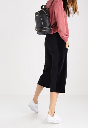 Sac à dos en cuir noir avec des accents dorés, forme arrondie, porté sur un chemisier rose et un pantalon large noir ; associé à des baskets blanches.