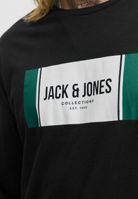 Schwarzes Langarm-T-Shirt mit einem grünen und weißen Logo-Rechteck, auf dem "JACK & JONES COLLECTIONS EST. 1990" in fetter Schrift steht. Weicher Stoff.