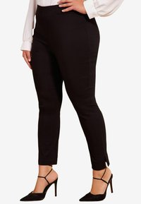 Schwarze, maßgeschneiderte Hosen aus einem glatten Stoff, mit schlankem Schnitt, Fronttaschen und Schlitzen am Knöchel, kombiniert mit hochhackigen Schuhen.
